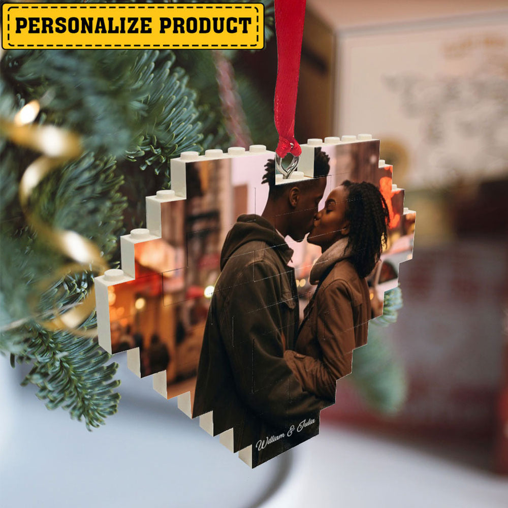 Custom photo brick heart ornament hanging from a glowing pine tree branch with a red ribbon and custom names at the base.