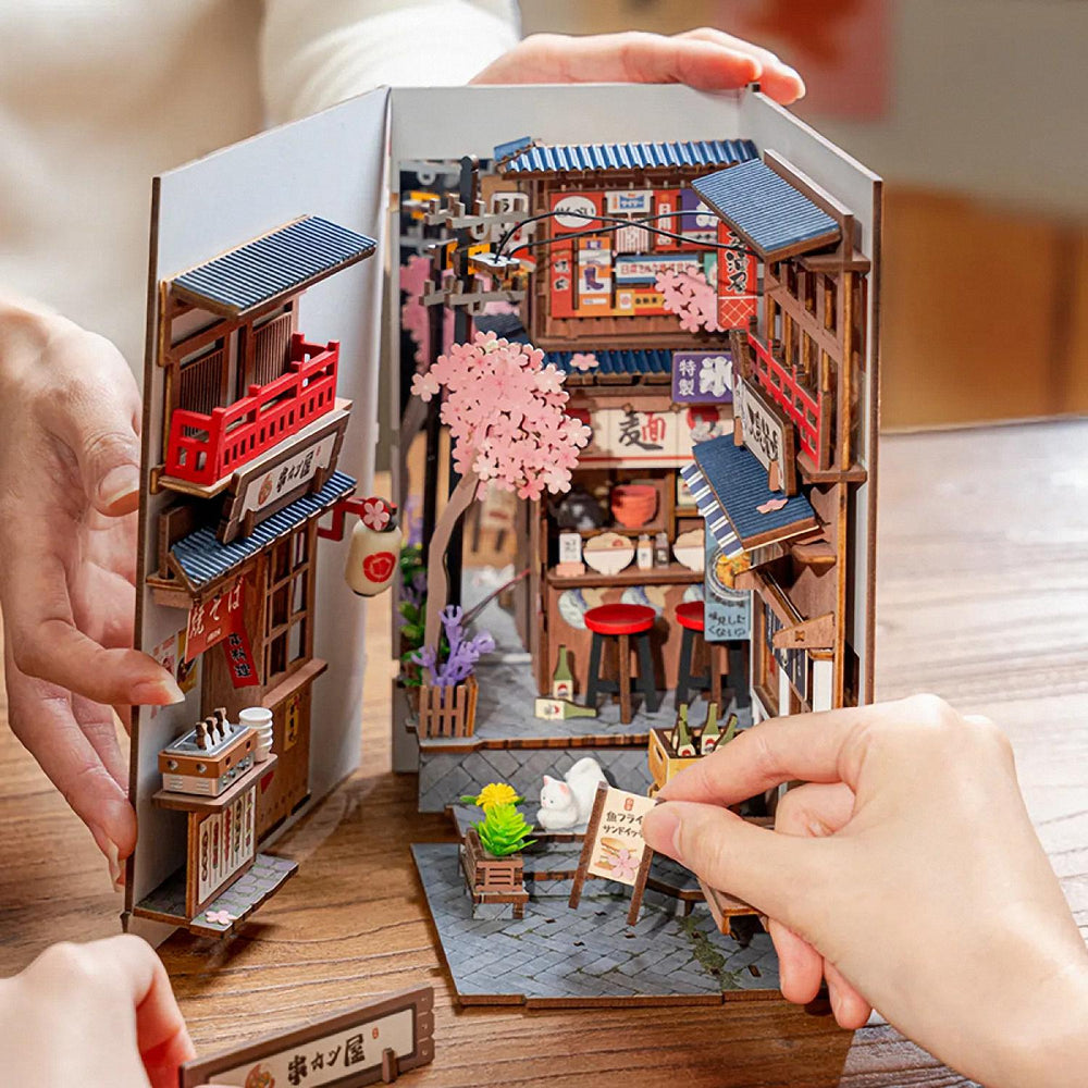 Hands assembling the pre-colored wooden pieces of the Sakura Wine Alley DIY book nook kit on a wooden table.