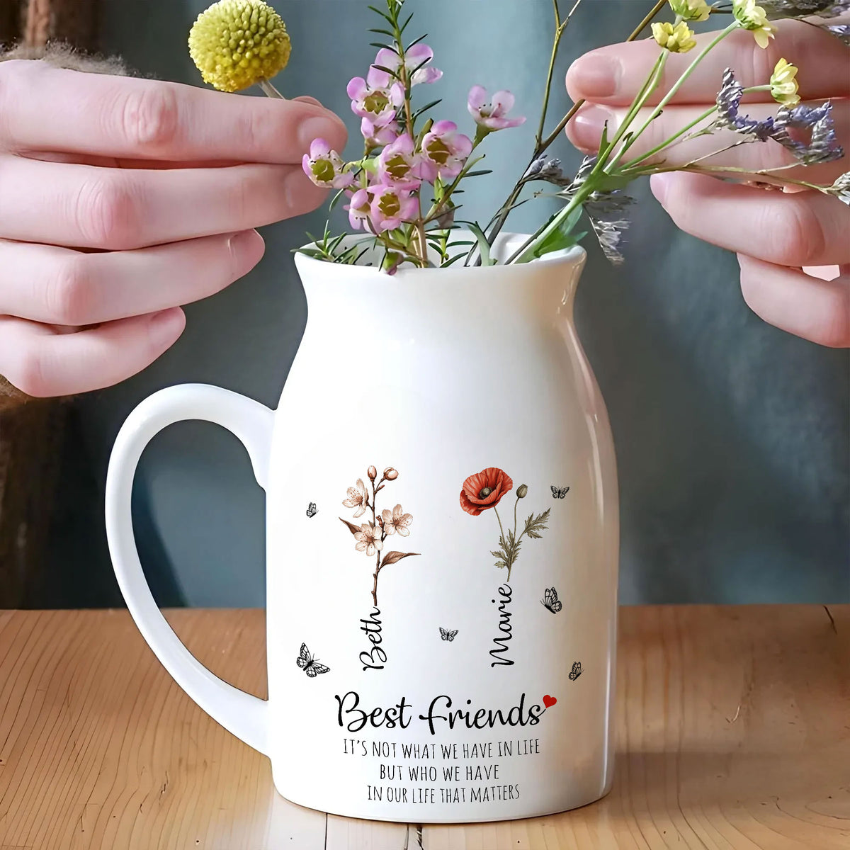 White ceramic flower vase with side handle on a wooden table, being filled with small colorful wildflowers by a pair of