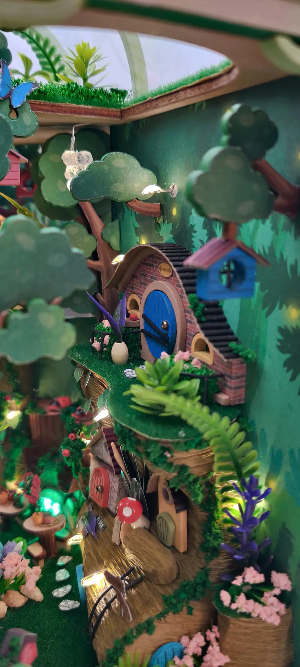 Close-up of the book nook puzzle showing a tiny brick cottage with a round blue door, green moss, and miniature plants.