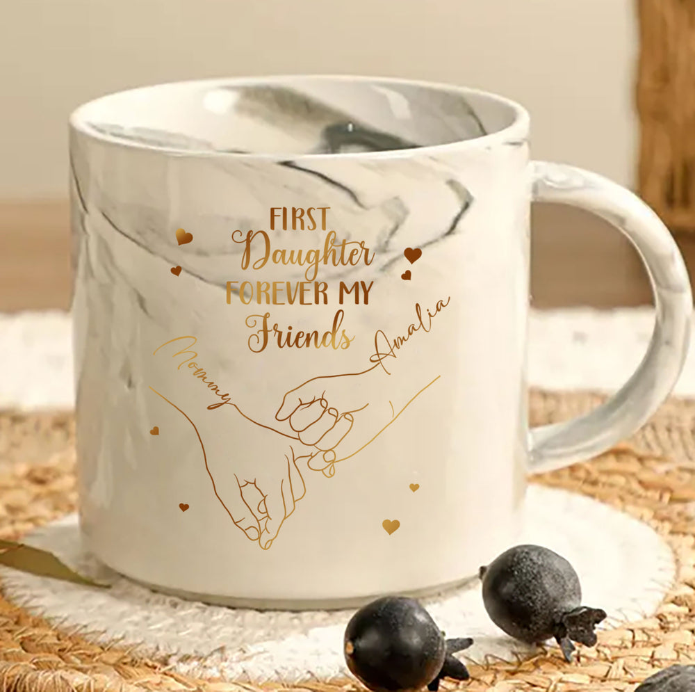 White marble mug with gold print showing pinky promise hands and the text First My Daughter Forever My Friend, on woven mat.