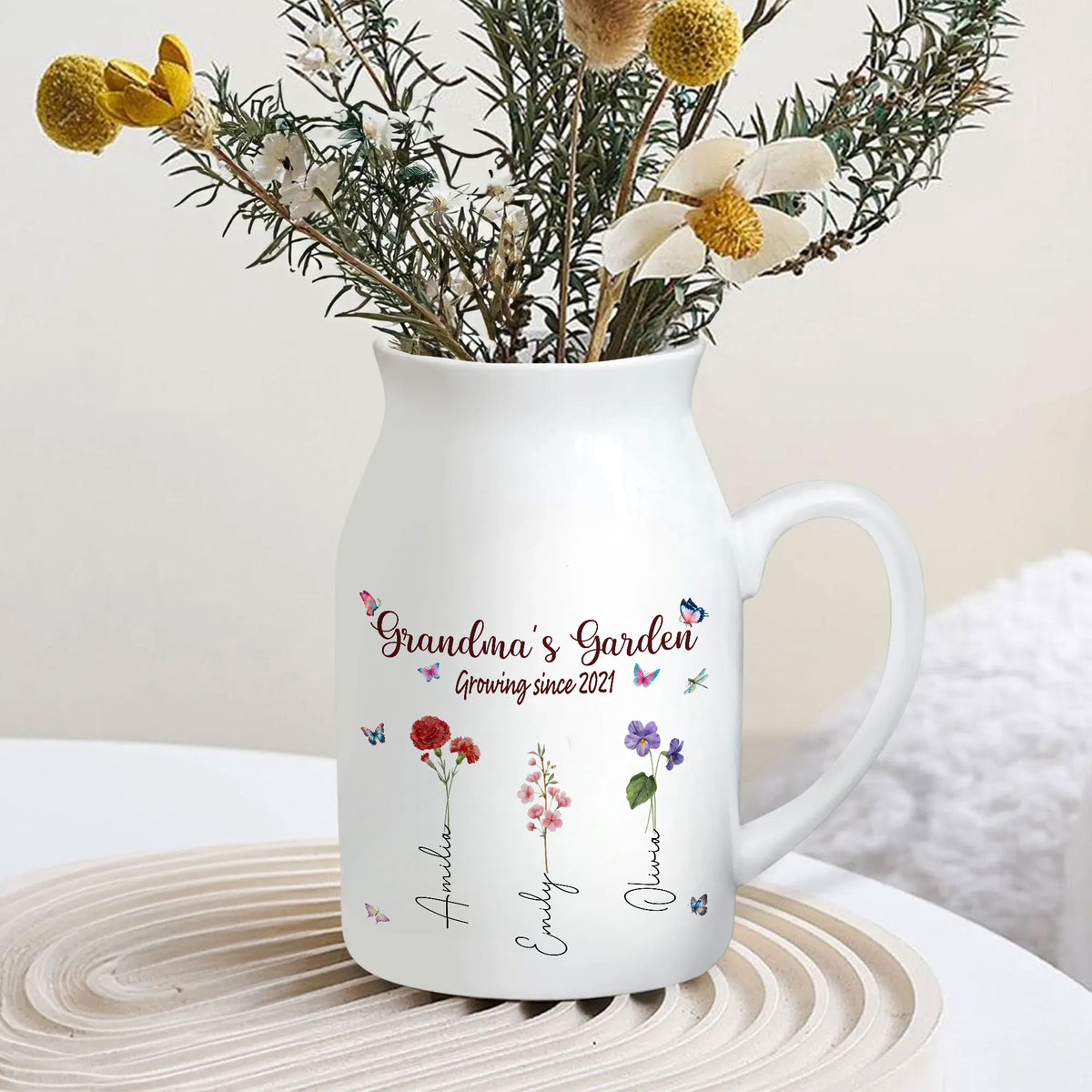 White ceramic flower vase filled with dried flowers, sitting on a round wooden tray in a bright room setting.