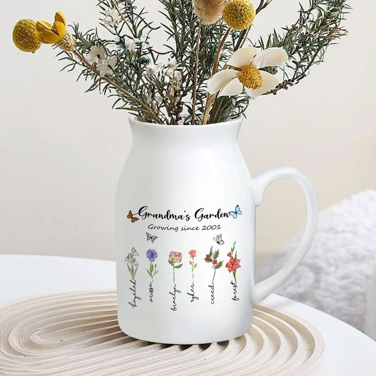 White ceramic flower vase with handle and colorful birth flowers design holding dried botanical stems on a table.