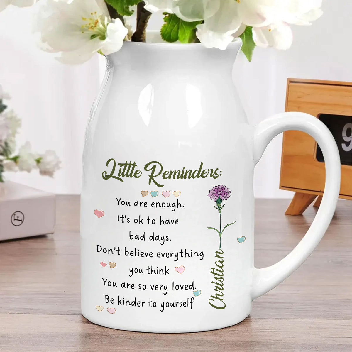 White ceramic flower vase holding white blossoms on a wooden tabletop, featuring printed motivational text, pastel hearts