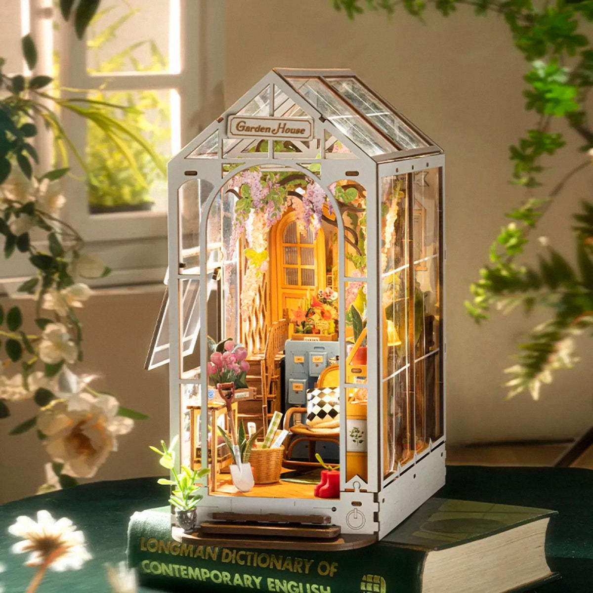 Miniature model of a greenhouse with interior details, placed on a book with plants around.