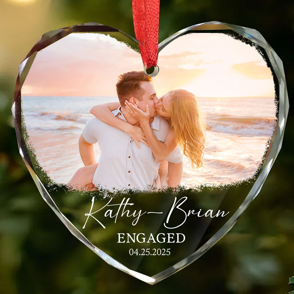 Close-up of a personalized heart-shaped glass ornament hanging with a red ribbon, showcasing a romantic beach engagement