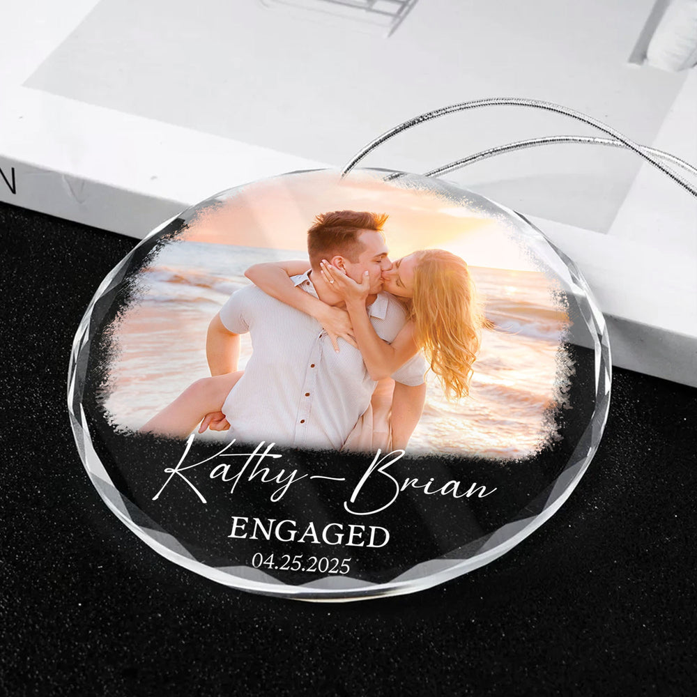 Custom photo glass ornament for engaged couple lying on a white surface with silver string attached.