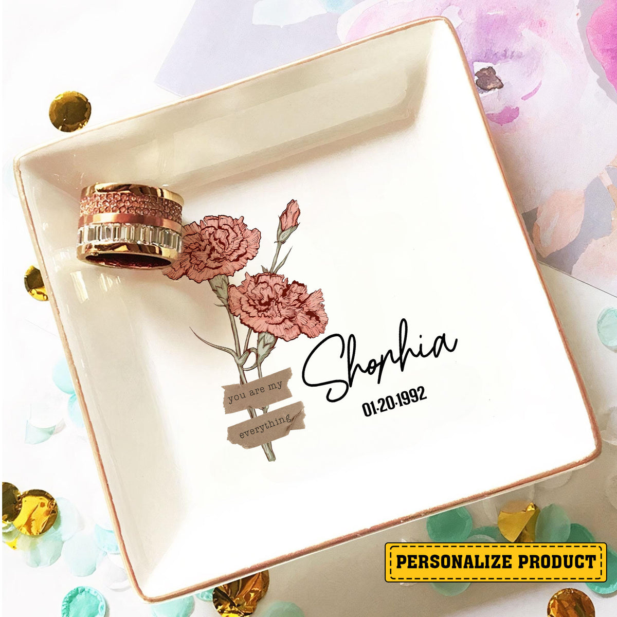 Personalized birth month flower ring dish holding a wide sparkling ring surrounded by gold confetti.