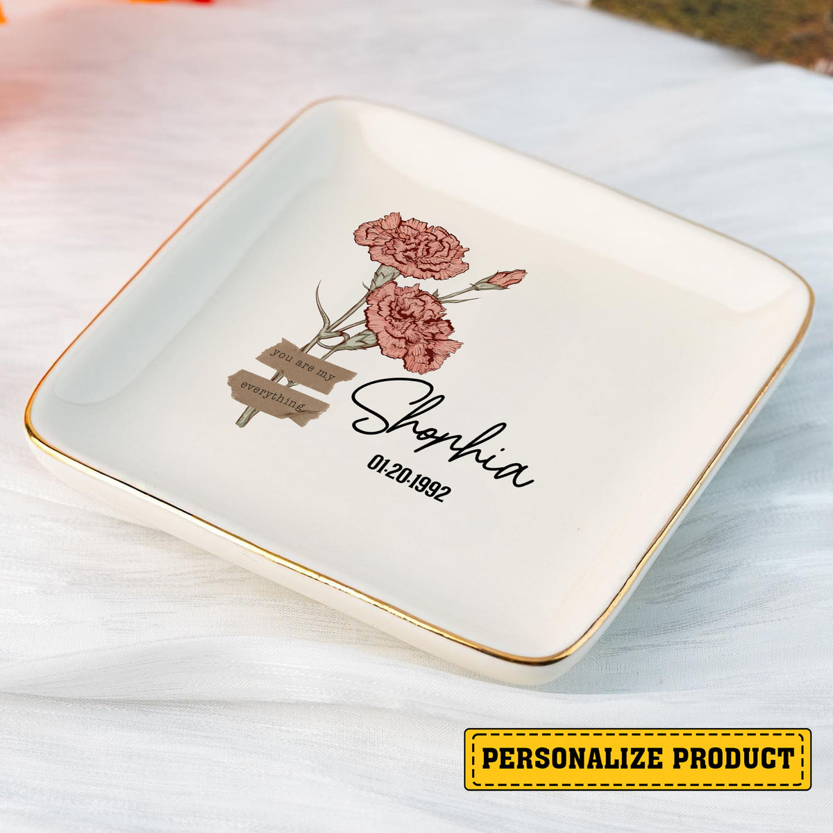 White square ring dish with pink carnations, gold rim, and custom script text on white fabric.