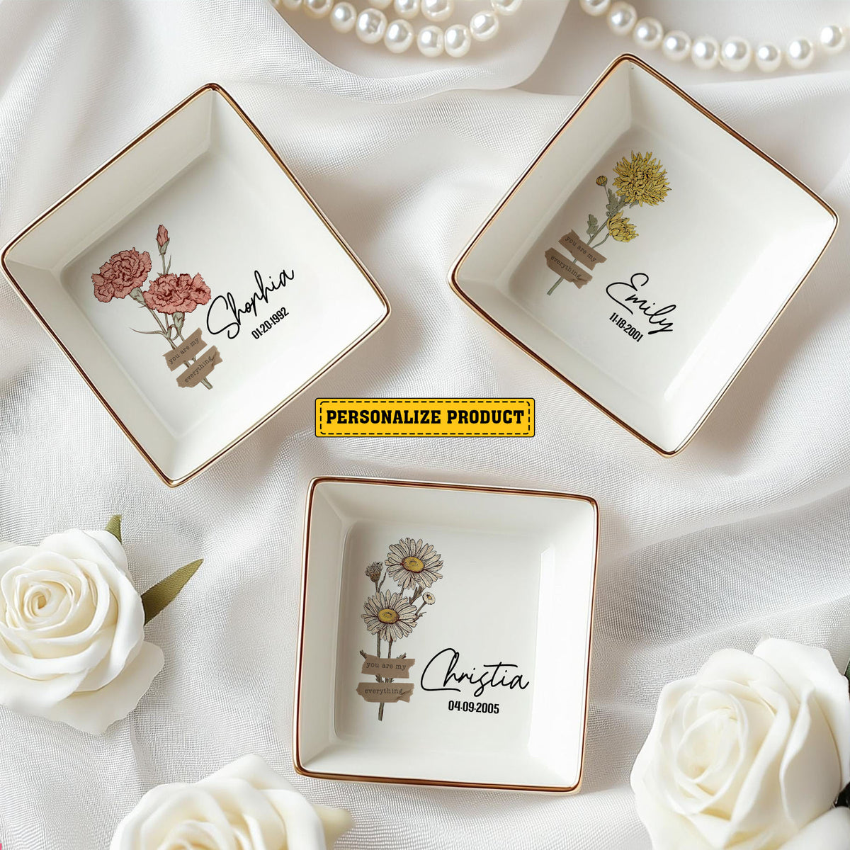 Three personalized birth month flower ring dishes with gold rims on white fabric with white roses.