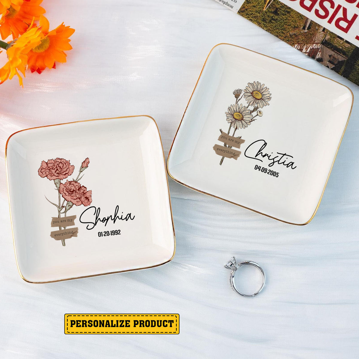 Two square white ceramic ring dishes with gold rims, featuring different birth month flower designs, names, and dates.