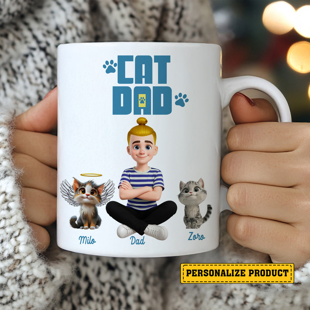 Close-up of hands holding a white ceramic mug printed with cartoon cat dad, angel cat, and living cat.