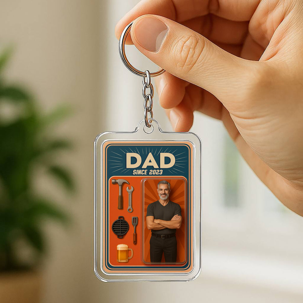 Hand holding personalized acrylic photo keychain featuring a man, tools, and beer graphics against a blurred indoor