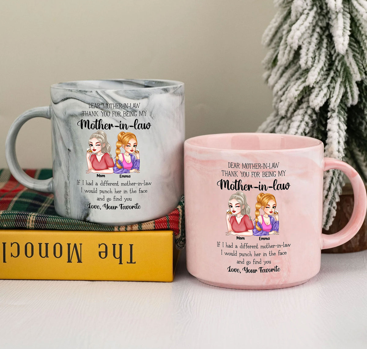 Grey and pink marble mugs featuring custom mother-in-law character design next to a miniature holiday tree.