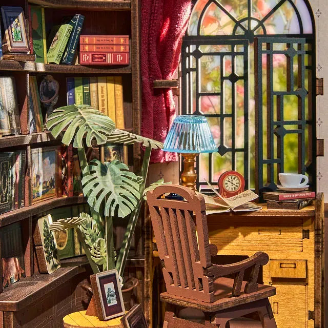 Close-up of the miniature library book nook interior showing a desk, rocking chair, potted plant, and arched window.